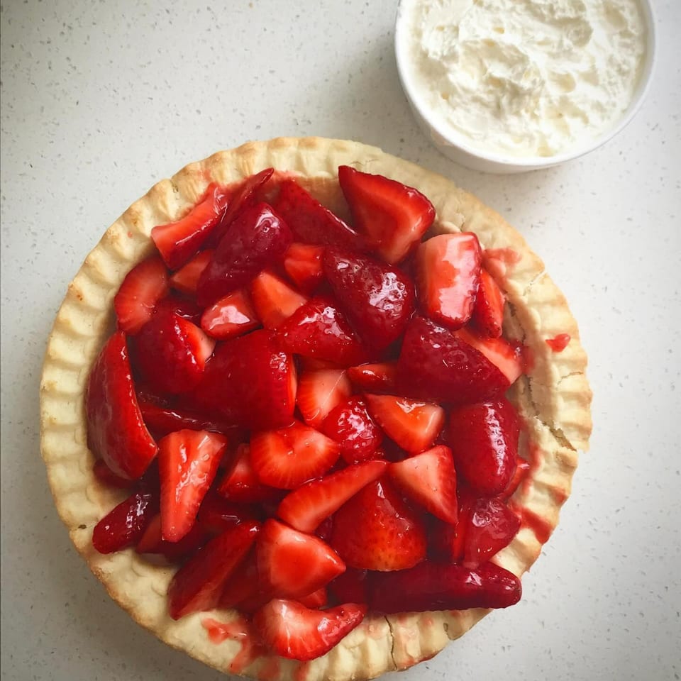 A strawberry pie with a flaky crust and a bowl of whipped cream on a white counter.