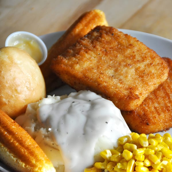 Fried chicken fillets with mashed potatoes, gravy, corn, a roll, and dipping sauce on a white plate.